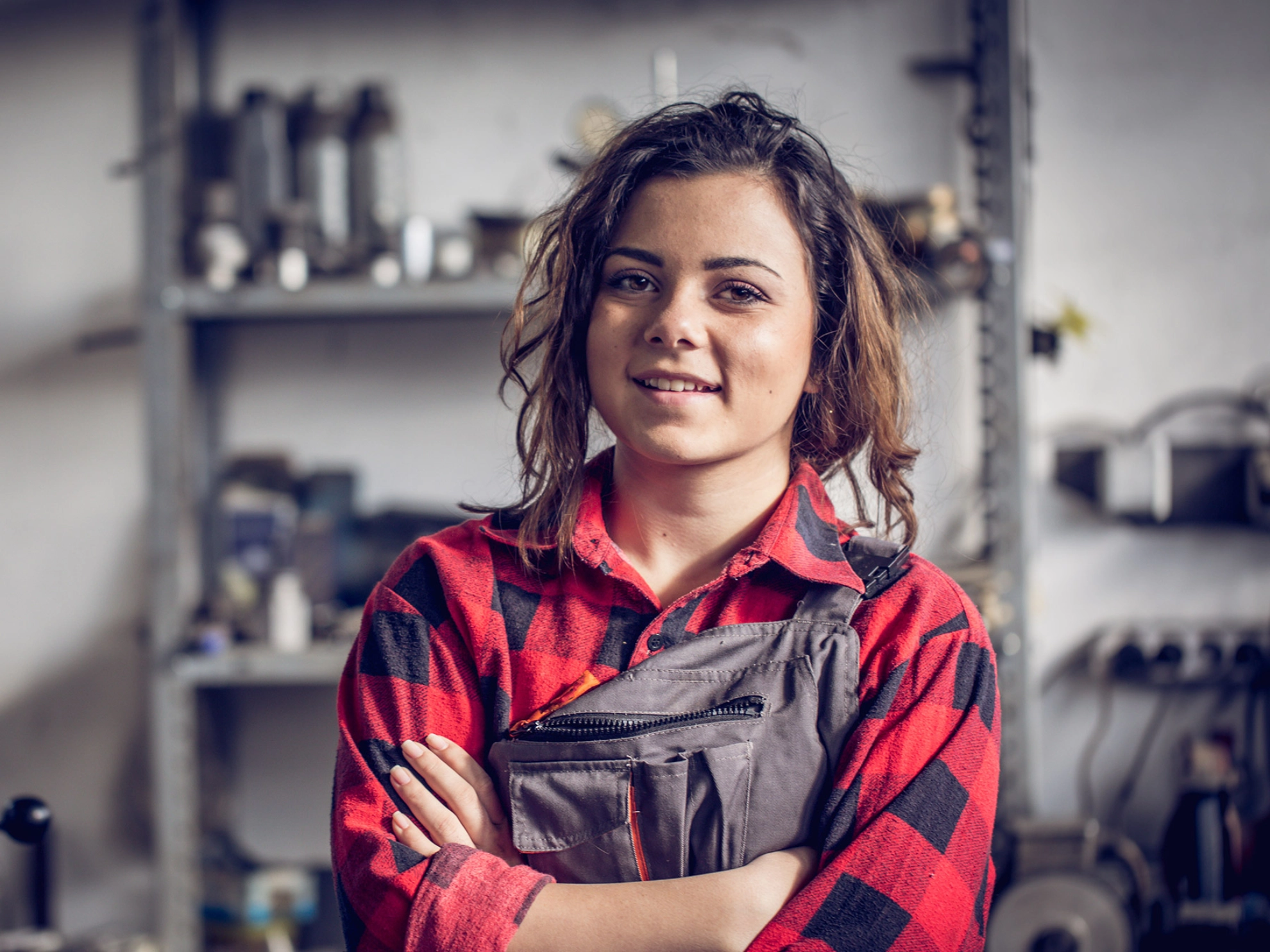 Junge Mechanikerin mit rotkariertem Hemd und Latzhose schaut mit verschränkten Armen selbstbewusst in die Kamera.
